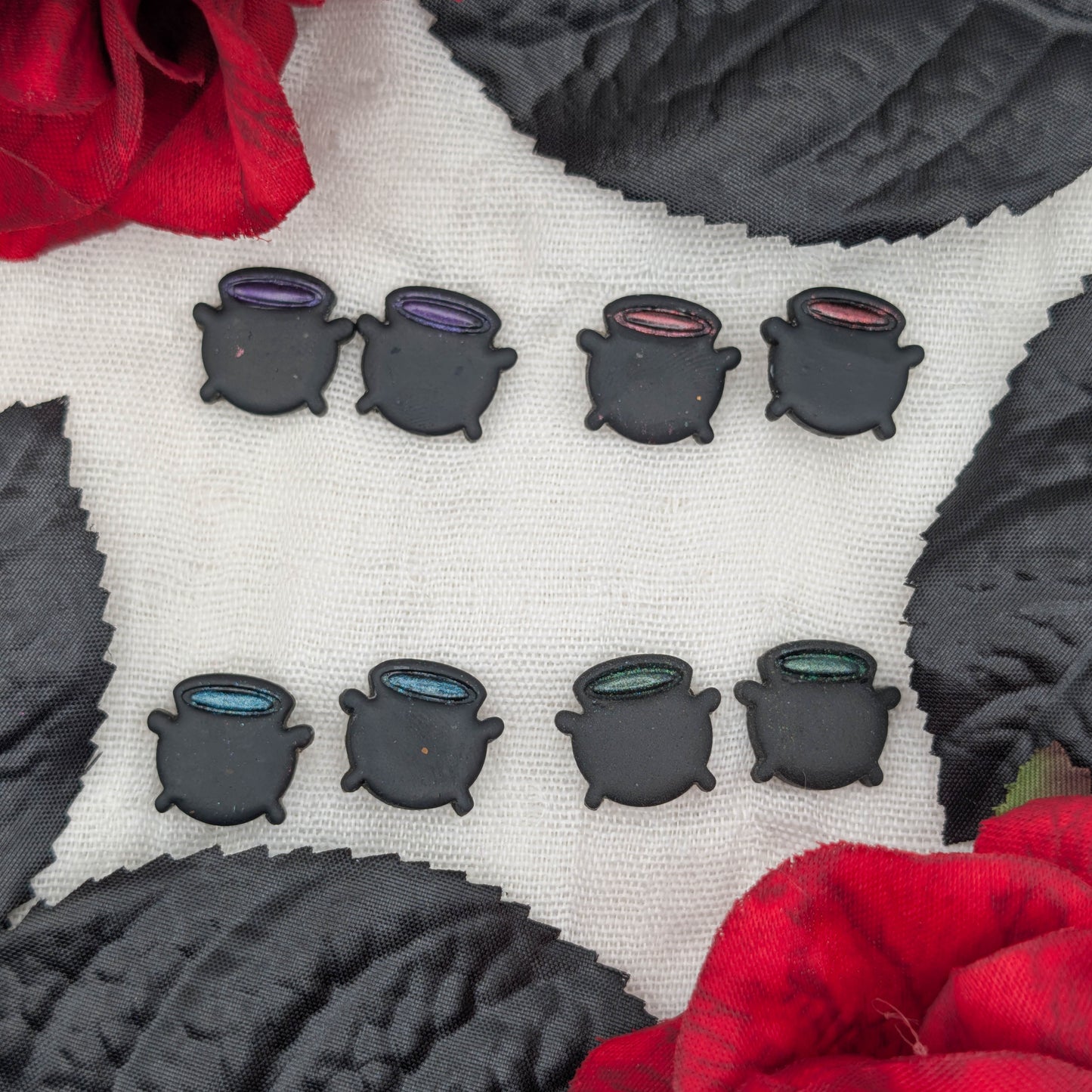 Eight small black cauldron-shaped accessories arranged in two rows on a white textured surface, surrounded by red roses and dark textured leaves. Each cauldron has a different colored rim: purple, blue, pink, green, and dark gray tones.