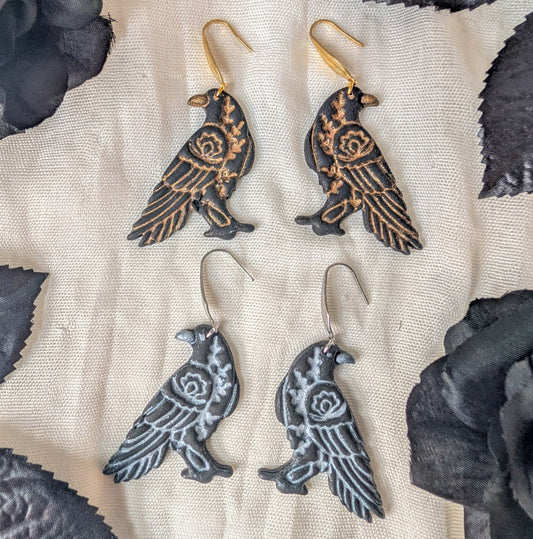 Gold and silver floral raven polymer clay earrings on cream fabric framed by black leaves