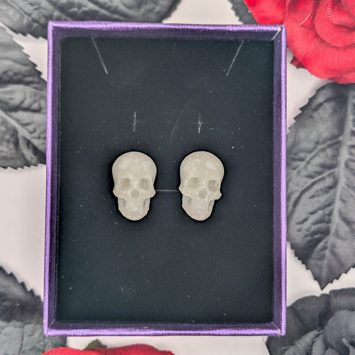 Glow-in-the-dark skull stud earrings displayed in a purple jewelry box with red and black flowers