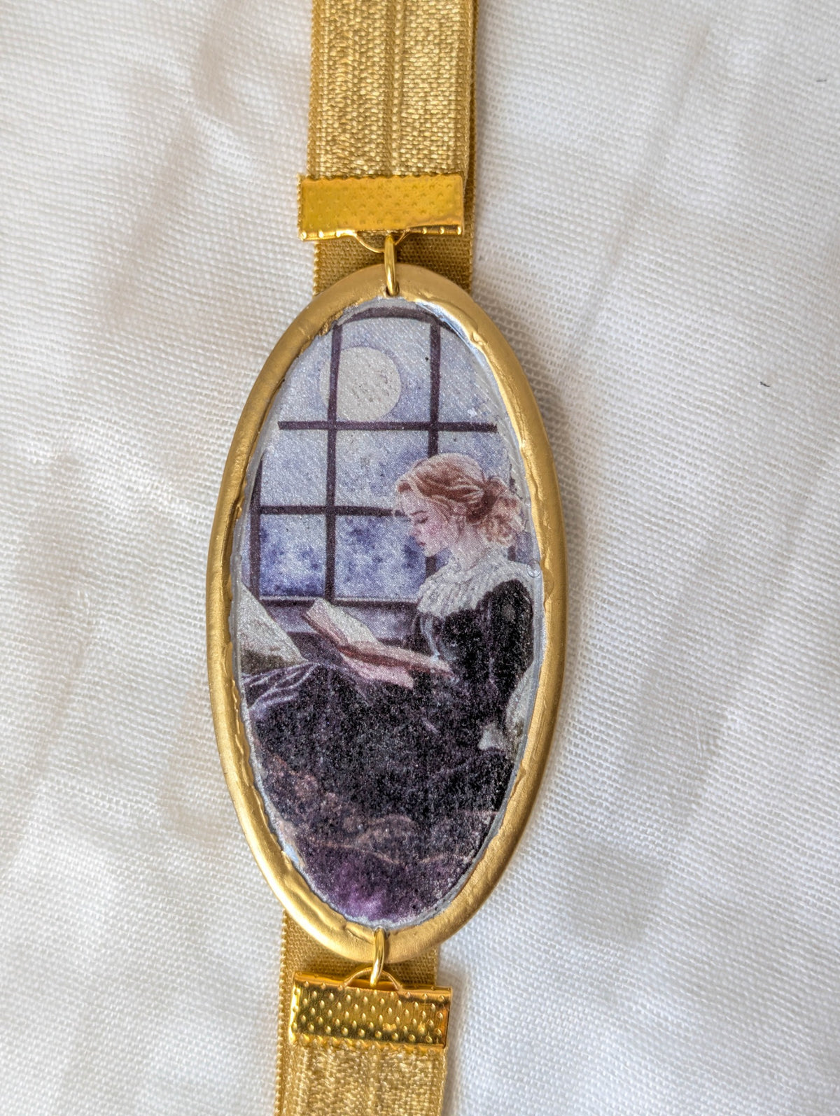 Close-up of gold framed bookmark showing a woman reading beside a moonlit window