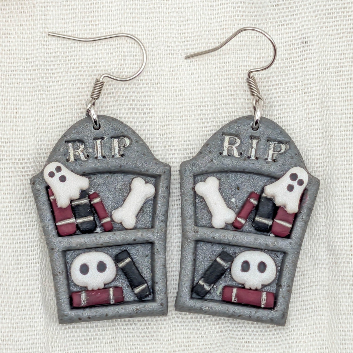 Close-up of polymer clay gravestone bookshelf earrings with burgundy and black books, ghosts, bones, and skulls