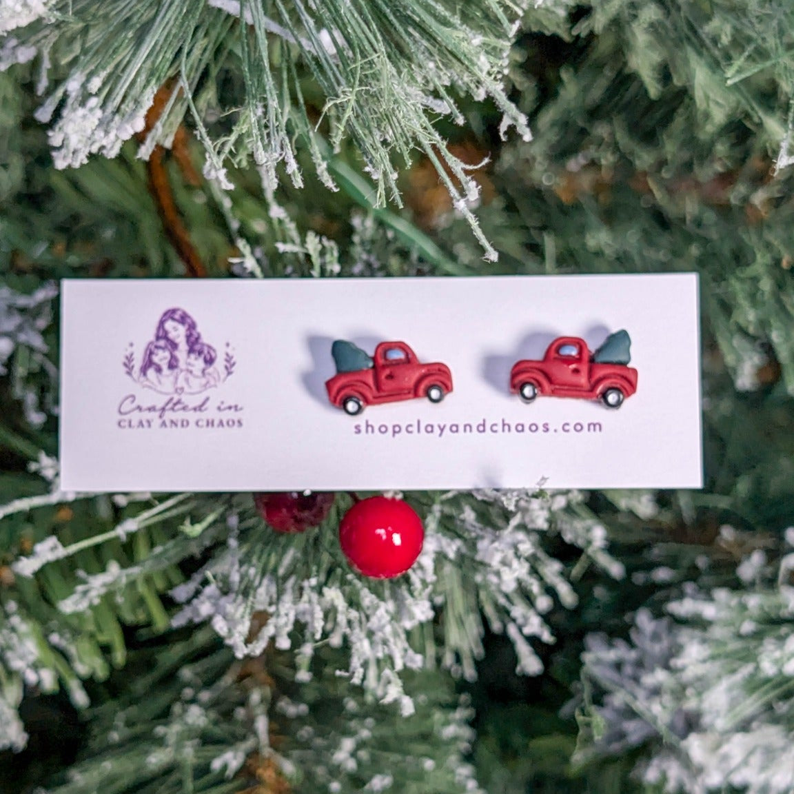 Mini red Christmas tree truck stud earrings displayed against snowy holiday greenery