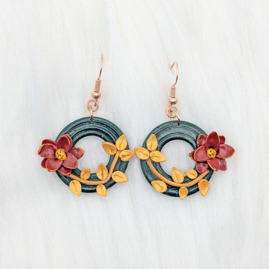 Two handmade poinsettia wreath dangle earrings with green wreaths, gold leaves, red flowers, and gold-plated stainless steel hooks.