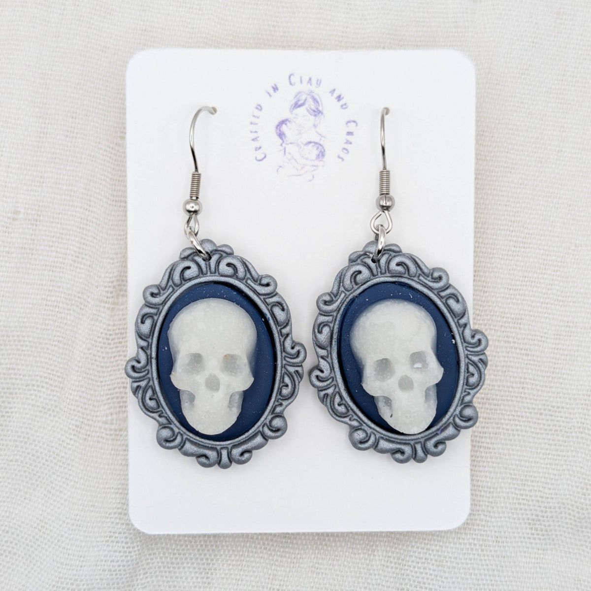 Silver oval vintage frame earrings with white glow-in-the-dark skulls, navy blue background, and surgical-grade stainless steel hooks