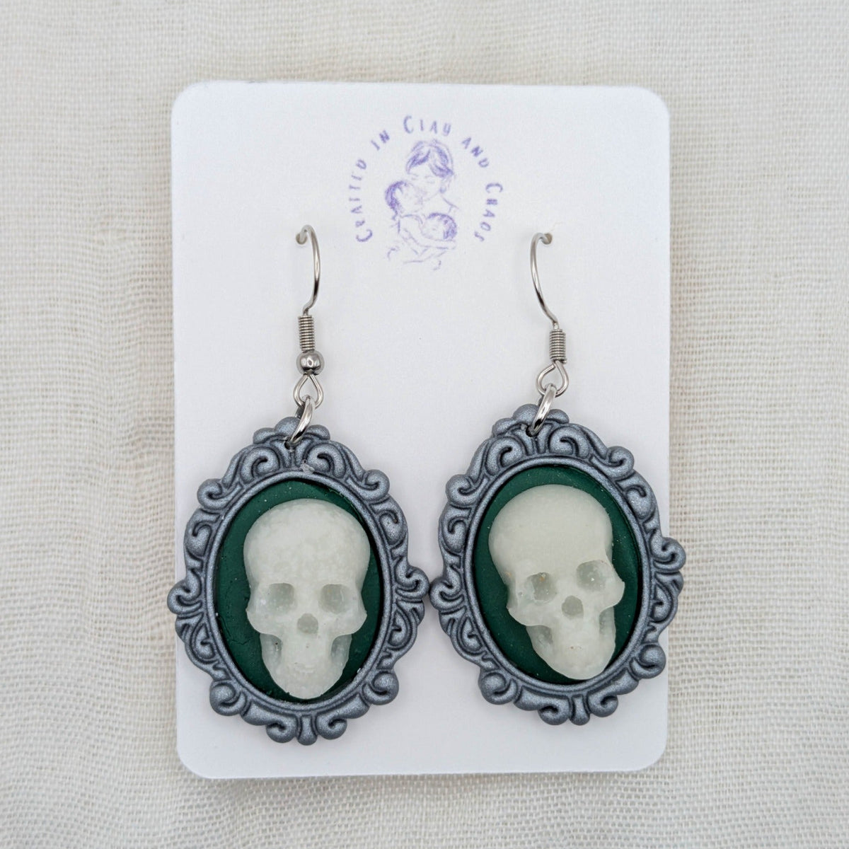 Silver oval vintage frame earrings with white glow-in-the-dark skulls, forest green background, and surgical-grade stainless steel hooks