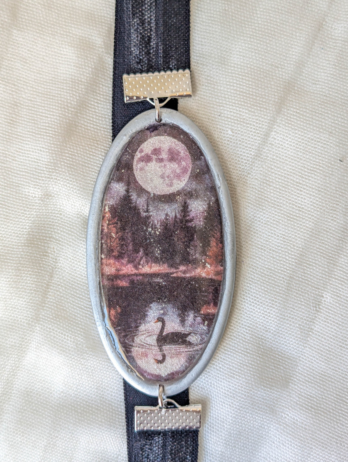 Close-up of silver framed bookmark showing a full moon, dark forest, reflective lake, and black swan