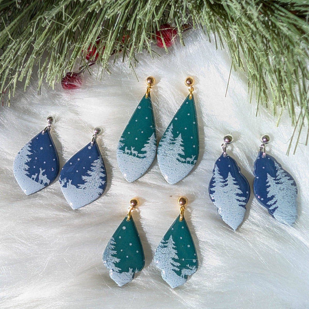 Blue and green snowy forest Christmas dangle earrings arranged beneath evergreen branches