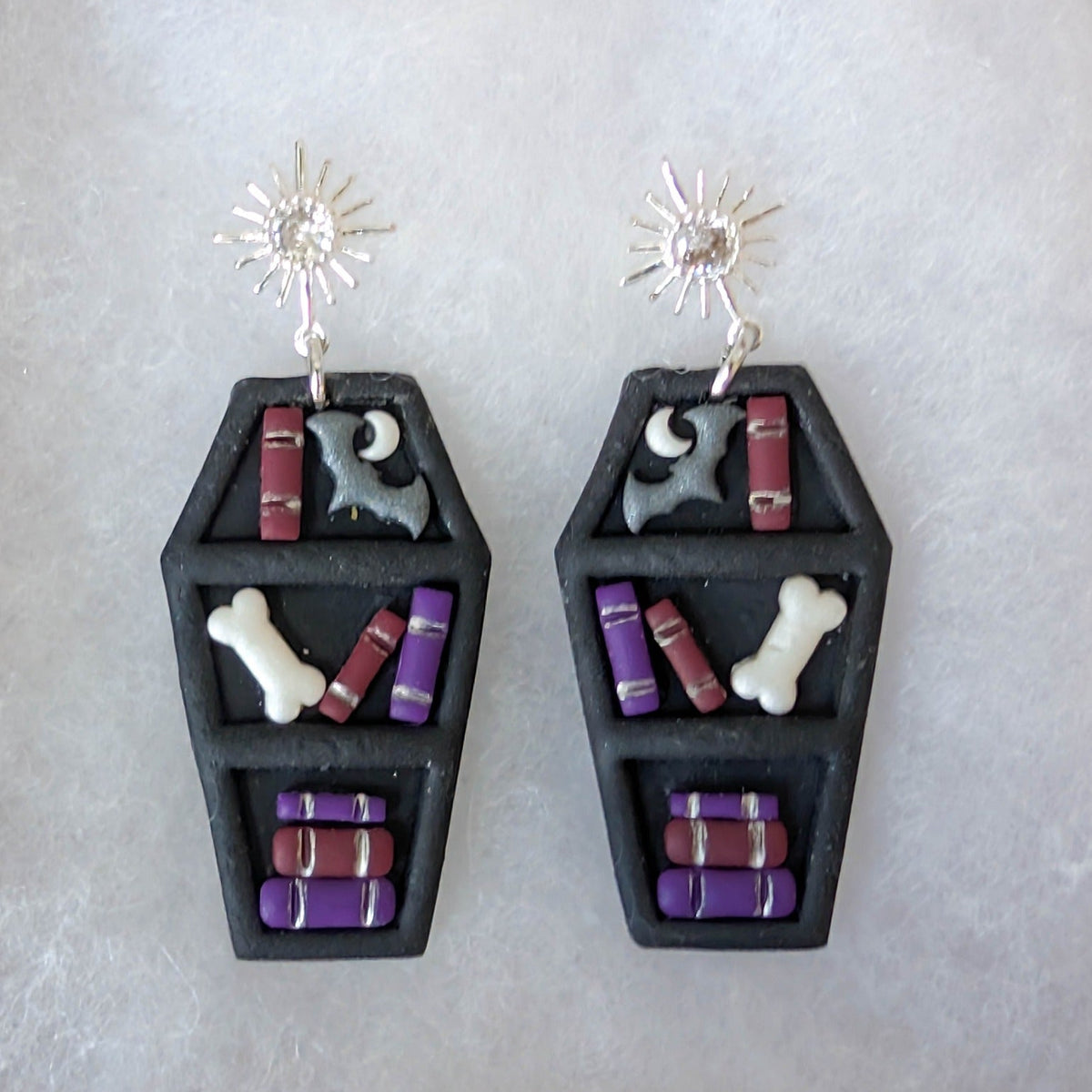 Close-up of black coffin bookcase earrings with burgundy and purple books, silver bats, crescent moons, and ivory bones